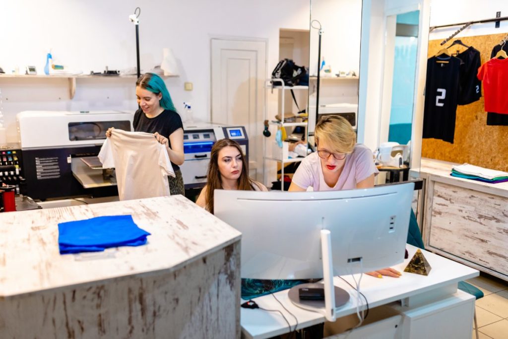 Trois femmes travaillant dans un atelier d'impression numérique textile, l'une tenant un t-shirt imprimé.